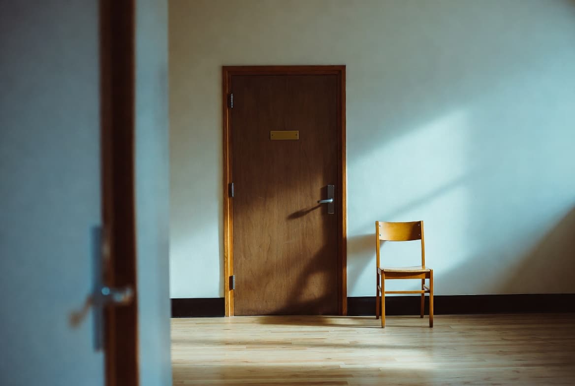 A closed wooden office door inside a school hallway in the afternoon, an empty wooden chair beside the door, sunlight slanting across a pale oak floor.